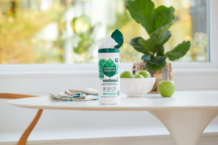 A container of Seventh Generation Multi-Surface Wipes with a garden mint scent is open on a white table beside folded towels and green apples. A leafy plant by a sunny window completes the fresh, clean atmosphere.