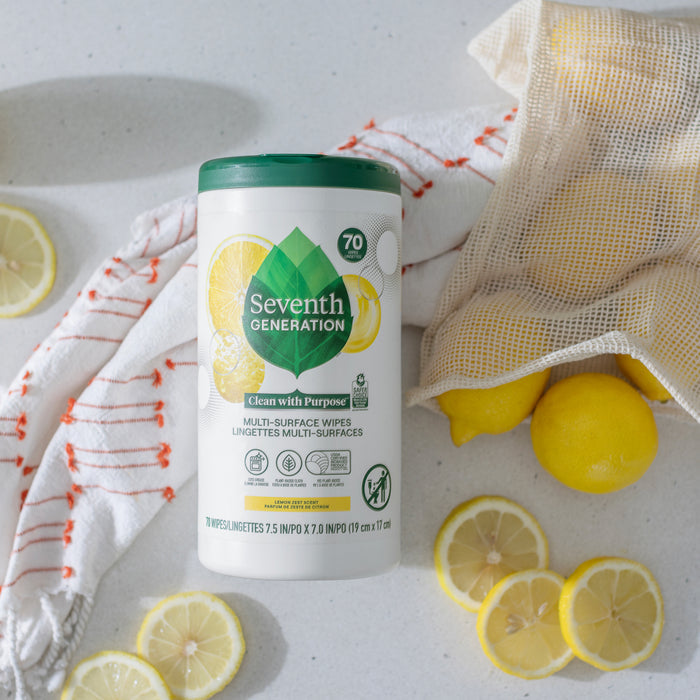 A container of Seventh Generation Multi-Surface Wipes, made from biobased materials, rests on the countertop with whole and sliced lemons around. Nearby is a reusable cloth bag brimming with lemons, while a striped towel subtly enters the scene.