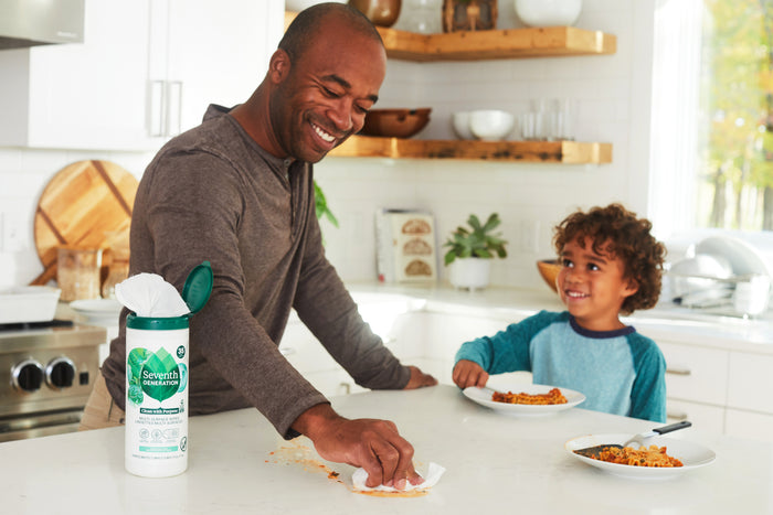 In a kitchen, a man uses Seventh Generation Multi-Surface Wipes to clean a counter spill. A curly-haired child smiles at the counter with food, while open shelves in the background display dishes, plants, and essential oils for decoration.
