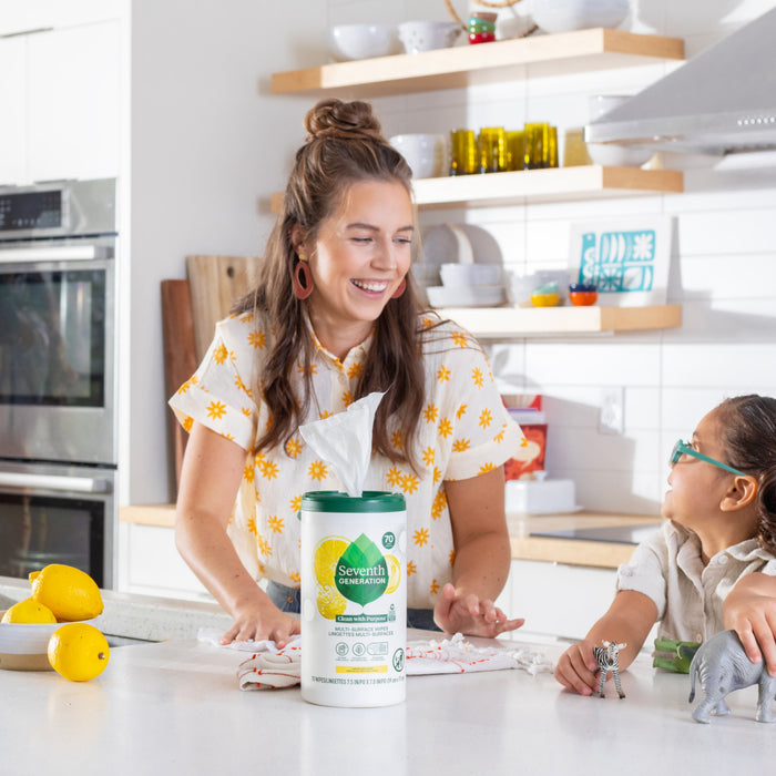 In a white kitchen, a woman in a floral blouse and a child in glasses share a warm moment. Multi-Surface Wipes by Seventh Generation sit on the counter, surrounded by lemons and small toy animals. She smiles warmly at him.