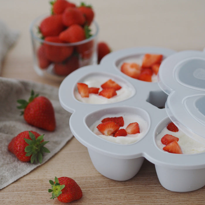The BEABA Multiportions™ Silicone Baby Food Freezer Tray, made from platinum-grade silicone, is filled with yogurt and topped with chopped strawberries. Fresh strawberries in a cup and some scattered on a wooden surface next to cloth hint at a summery, fresh dessert preparation.