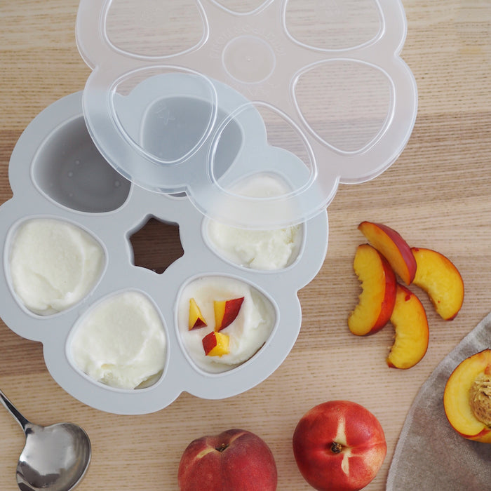 A BEABA Multiportions™ Silicone Baby Food Freezer Tray filled with yogurt and diced peaches sits invitingly with its lid slightly open. Nearby, whole and sliced peaches accompany a spoon, ideal for baby food storage or as a Babycook accessory.