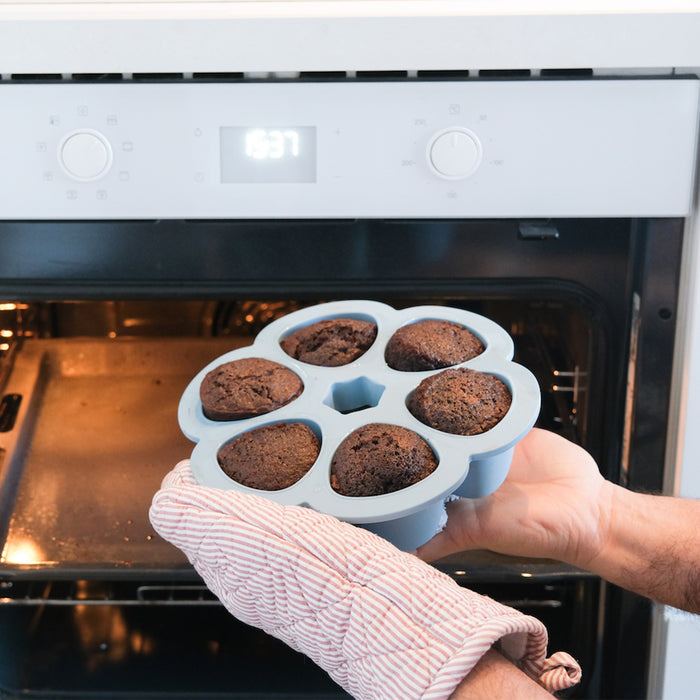 Hands wearing oven mitts hold a BEABA Multiportions™ Silicone Baby Food Freezer Tray filled with six baked chocolate muffins. The oven door is open, and the digital clock shows 6:37.