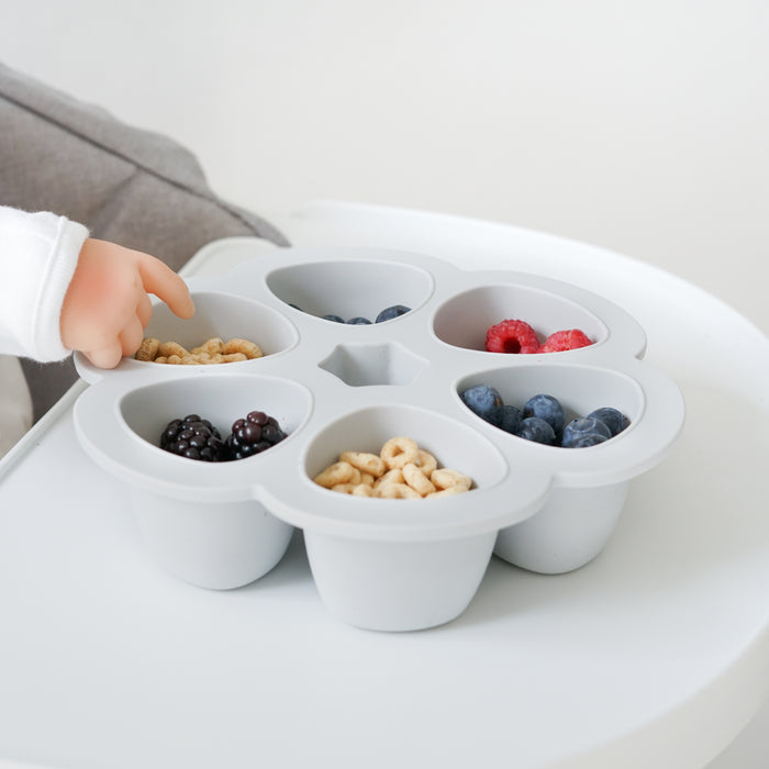 A childs hand reaches for the BEABA Multiportions™ Silicone Baby Food Freezer Tray, neatly divided with cereal, raspberries, blueberries, blackberries, and nuts. This perfect Babycook complement sits on a white table beside a gray fabric chair.