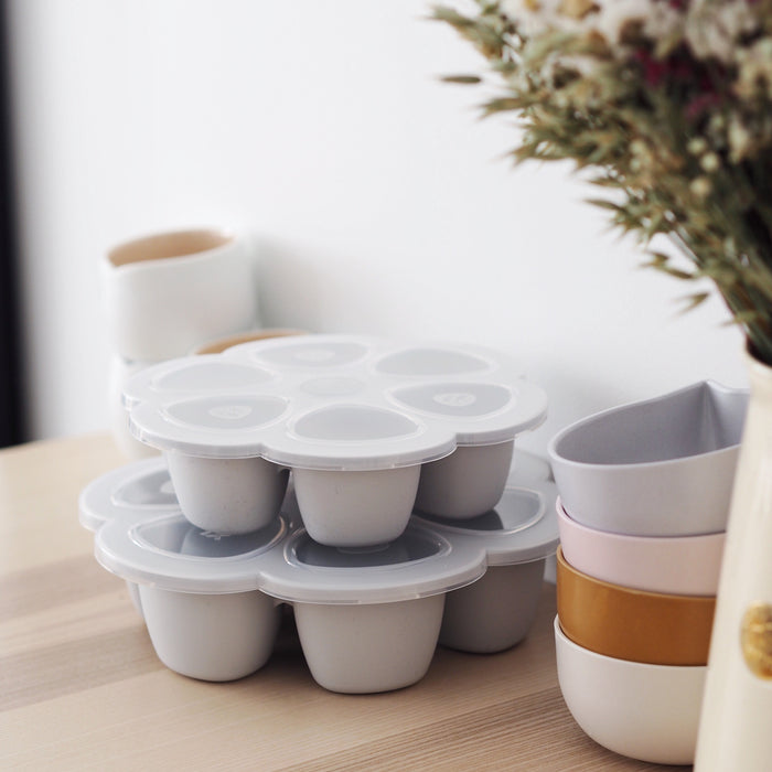 BEABAs Multiportions™ Silicone Baby Food Freezer Trays, made from platinum-grade silicone, are stacked on a wooden surface next to pastel-colored bowls. Blurred dried flowers add an aesthetic touch, and these trays are ideal for baby food storage or as a Babycook complement.