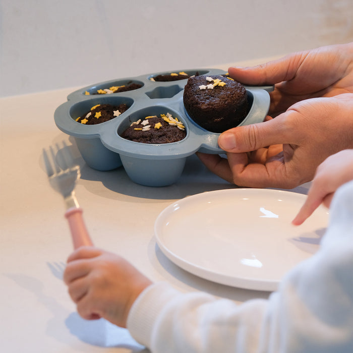 A person holds a BEABA Multiportions™ Silicone Baby Food Freezer Tray, platinum grade, containing six heart-shaped chocolate muffins with star sprinkles. Nearby, a child eagerly reaches with a fork towards a white plate on the table.