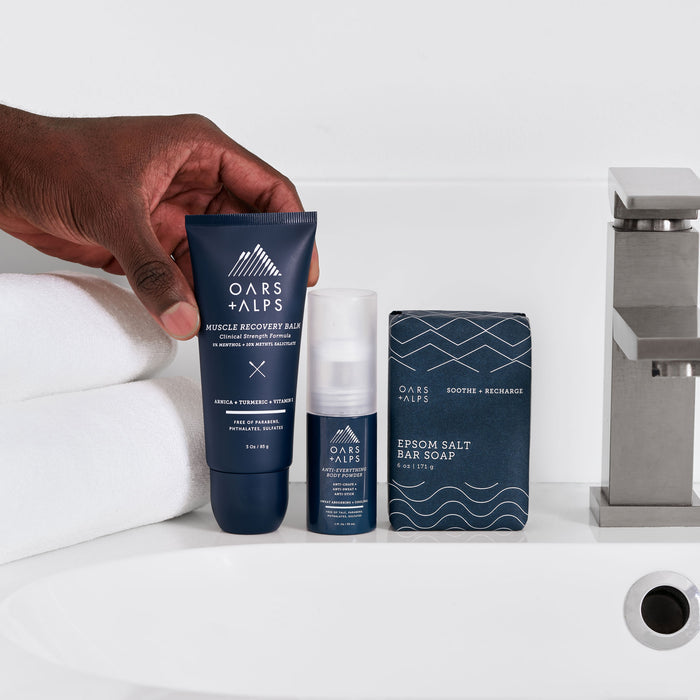 A hand holds a tube of Oars + Alps Muscle Recovery Balm for sore muscles by a bathroom sink, with folded white towels, a modern faucet, and other Oars + Alps products like deodorant and Epsom Salt Bar Soap in the background.