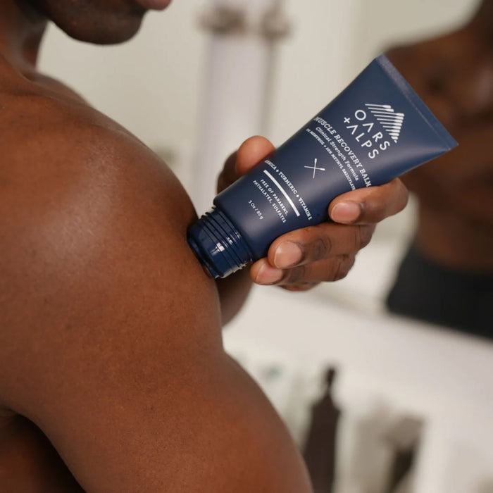 In a bathroom, someone applies Oars + Alps Muscle Recovery Balm to their shoulder. The blue tube, labeled Muscle Recovery Balm, is held near the skin and features muscle-soothing ingredients.