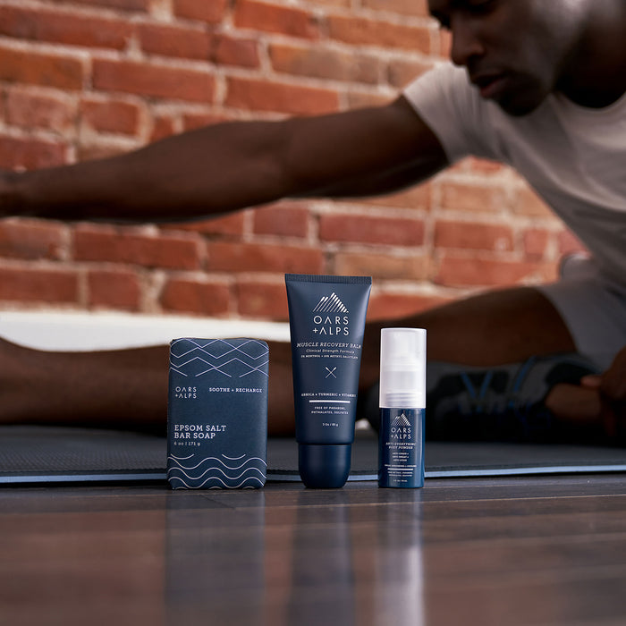 Three Oars + Alps skincare products, including their Muscle Recovery Balm, are arranged on a yoga mat in the foreground, as a person stretches on the mat before a brick wall in the background.