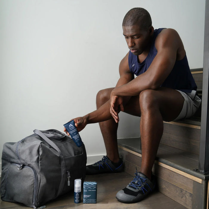 A man in athletic wear sits on wooden stairs, holding a blue bottle as Oars + Alps Muscle Recovery Balm and skincare products rest nearby, ready for use before or after his workout.