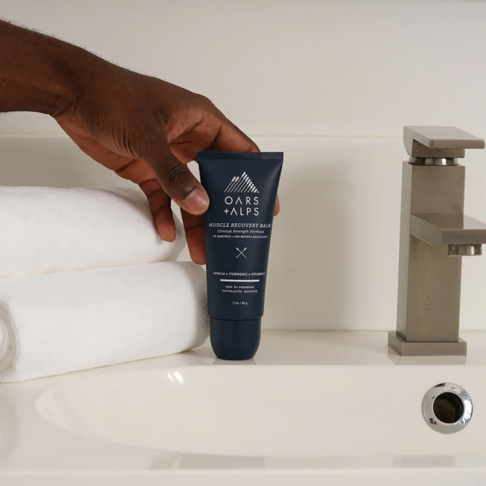A hand holds a tube of Oars + Alps Muscle Recovery Balm above a white sink, beside two rolled white towels and a silver faucet.
