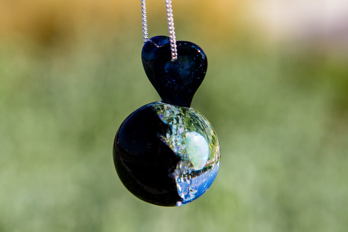 Flame worked glass pendant with a heart shaped top and a spherical body displaying a white heart opal and cremains against a colorful dichroic background