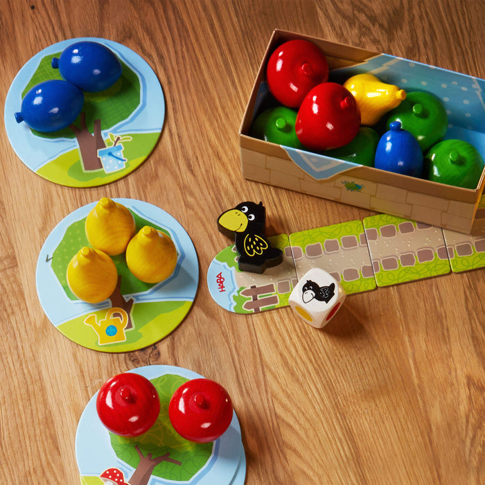 Vibrant apple-shaped pieces in red, blue, green, and yellow sit on tree-patterned mats. The First Orchard Game box from HABA is joined by a black crow figure and a die with fruit images, making this cooperative game ideal for early learning on any wooden surface.