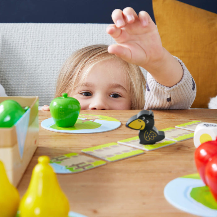 A blonde child peeks over a table, reaching for a green apple toy in HABAs My Very First Games - First Orchard, a delightful board game filled with fruit-shaped pieces and featuring a crow figurine, ideal for cooperative play.