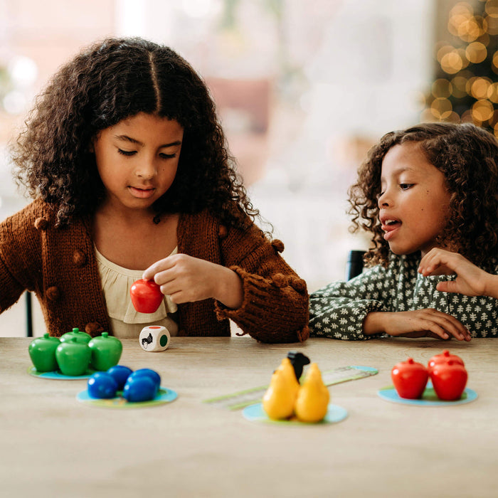 Two curly-haired girls sit at a table playing HABAs My Very First Games - First Orchard, a cooperative game with colorful plastic fruits. The girl on the left, in a brown cardigan, holds a red apple as her friend in a green patterned top watches—ideal for early learning fun.