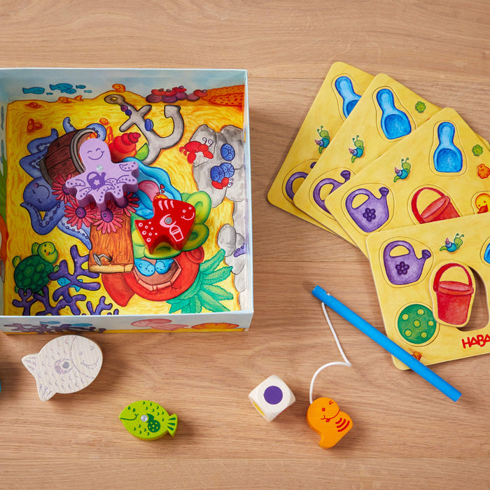 On a wooden table sits HABAs My Very First Games - Here, Fishy, Fishy! Magnetic Game, a colorful childrens board game including a blue box with underwater illustrations, fish-shaped pieces, a fishing rod for hand-eye coordination, dice, and yellow cards with watering cans to enhance early development skills.