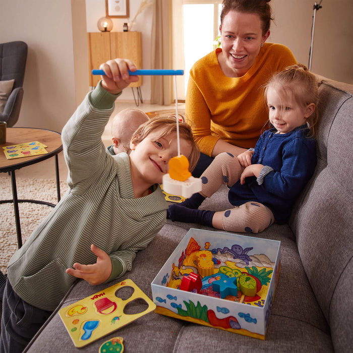 A young child smiles at the camera while playing with HABAs My Very First Games - Here, Fishy, Fishy! Magnetic Game, honing hand-eye coordination. Another child and an adult watch from a gray sofa. A colorful puzzle game adorns the table in the cozy, well-lit room.
