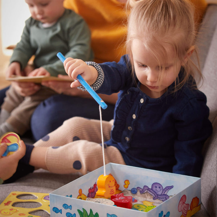 On a gray sofa, a young girl in a navy dress enhances her hand-eye coordination with HABAs My Very First Games - Here, Fishy, Fishy! Magnetic Game, using a blue rod to catch colorful wooden fish from a box. Another child nearby focuses on reading.
