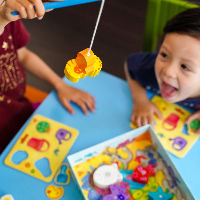 Two children energetically play with a wooden puzzle at a blue table. One child is engaged with HABAs My Very First Games - Here, Fishy, Fishy! Magnetic Game, holding colorful animal pieces on a string. The other looks excited, while a puzzle box containing various shapes sits on the table.