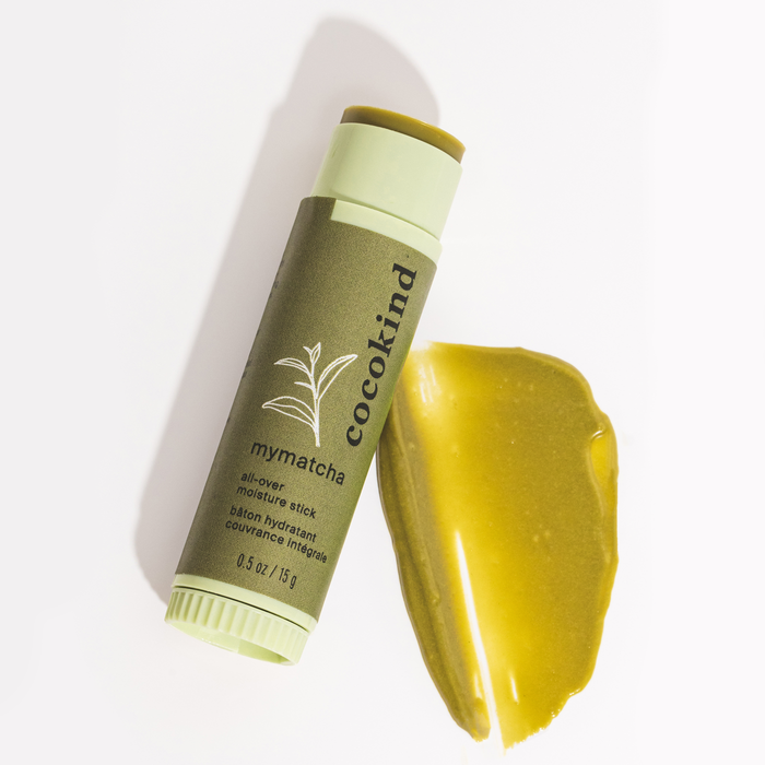 A green MyMatcha All-Over Moisture Stick by Cocokind is on a white background, beside a creamy smear. Perfect for dark circles and dry lips, it features plant artwork on the packaging.