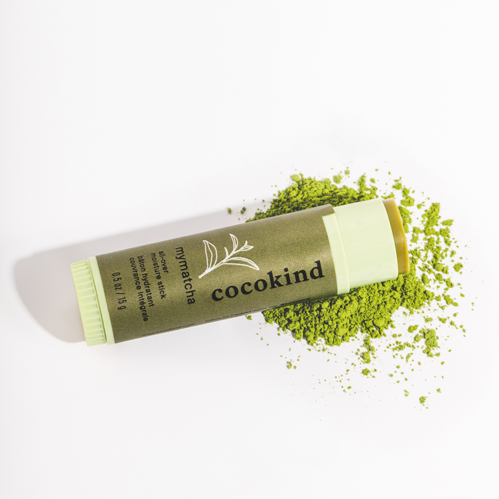 The Cocokind MyMatcha All-Over Moisture Stick rests on its side amidst scattered matcha powder, highlighting its leaf-designed packaging and product details on a white background. Ideal for addressing dark circles or hydrating dry lips.