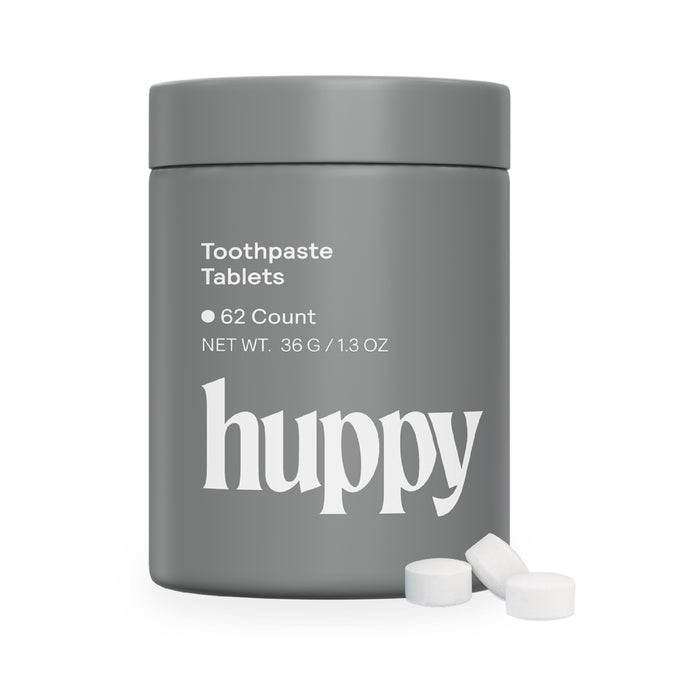 A gray huppy tin labeled “Toothpaste Tablets with Tin, 62 count, Net Wt. 36 g / 1.3 oz” holds sustainable toothpaste tablets. Three white cavity-fighting tablets are placed next to the container.