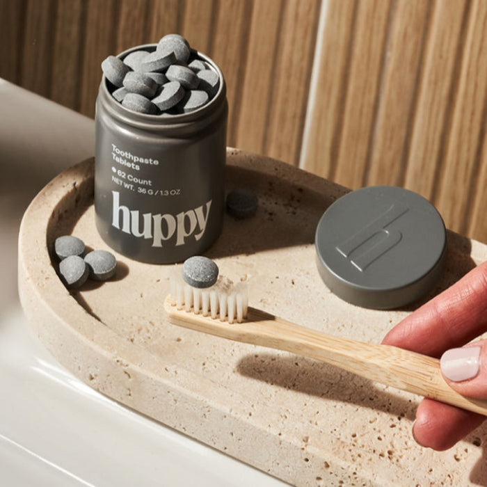 A hand holds a bamboo toothbrush beside Huppy Toothpaste Tablets with Tin—sustainable toothpaste tablets—displayed on a beige stone tray, with several tablets scattered around.