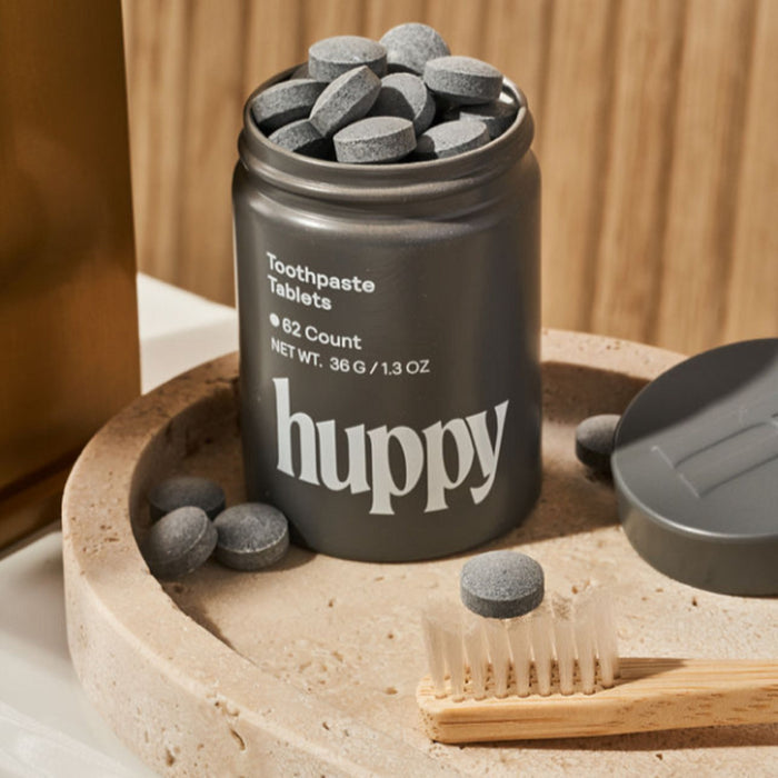 A tin labeled Huppy holds sustainable Toothpaste Tablets, some scattered nearby. A wooden toothbrush with beige bristles rests on a round beige tray, while the tin lid sits to the side.