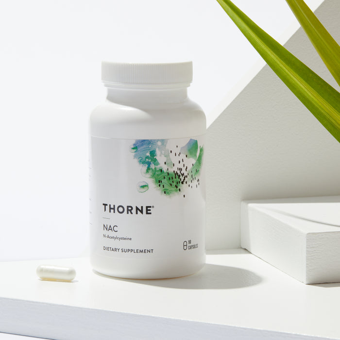 A white bottle of Thornes NAC - N-Acetylcysteine rests on a sunlit white surface. The label, featuring abstract green and blue artwork, suggests antioxidant properties. A single capsule and a touch of green plant add tranquility to the scene.