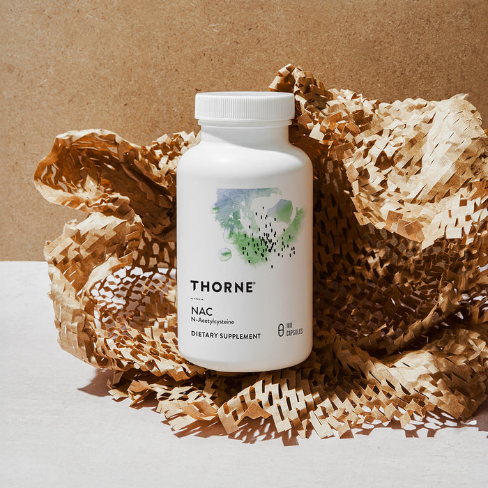A white bottle of Thorne NAC - N-Acetylcysteine dietary supplement, recognized for supporting liver and immune health, rests on crinkled brown paper with a beige background. The label shows a green and blue abstract pattern.