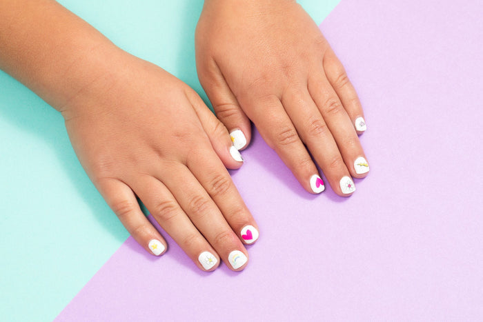 A childs nails are painted white and decorated with Terra Beauty Bars Nail Art Stickers, featuring hearts, stars, and circles. Set against a pastel blue and purple backdrop, this playful design embraces sustainable nail care ideals.