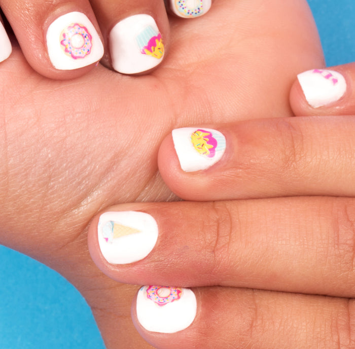 Close-up of hands with white nail polish and Terra Beauty Bars Nail Art Stickers featuring cupcakes and ice cream cones on a bright blue background, offering a delightful and sustainable nail care experience.