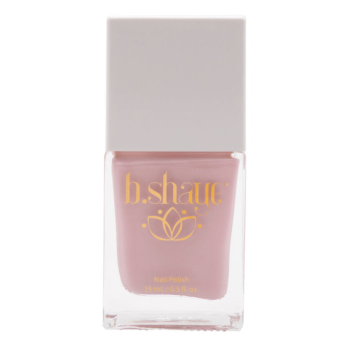 A 15 ml (0.5 fl. oz.) bottle of non-toxic nail polish with pink color is inside a clear container, topped with a square, light gray cap. The label features the brand b.shaye in gold text and a floral design, showcasing this clean beauty product.