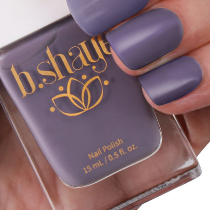 A hand holds a bottle of b.shaye Nail Polish in matte lavender. The vegan, cruelty-free polish features gold lettering and a stylized flower logo on its clear bottle, embodying clean beauty.