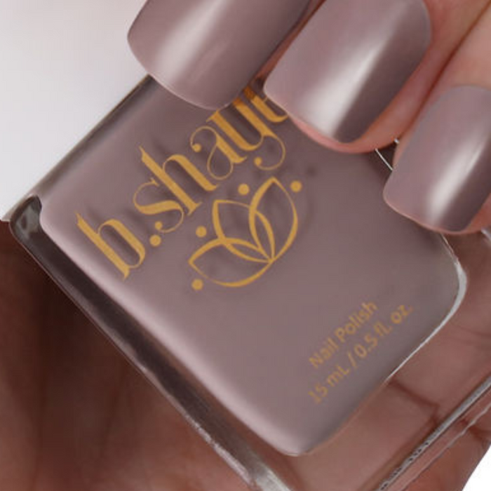 A hand holds a bottle of b.shaye Nail Polish in mauve, with matching painted nails. The polish is non-toxic, vegan, and cruelty-free, part of a clean beauty range.