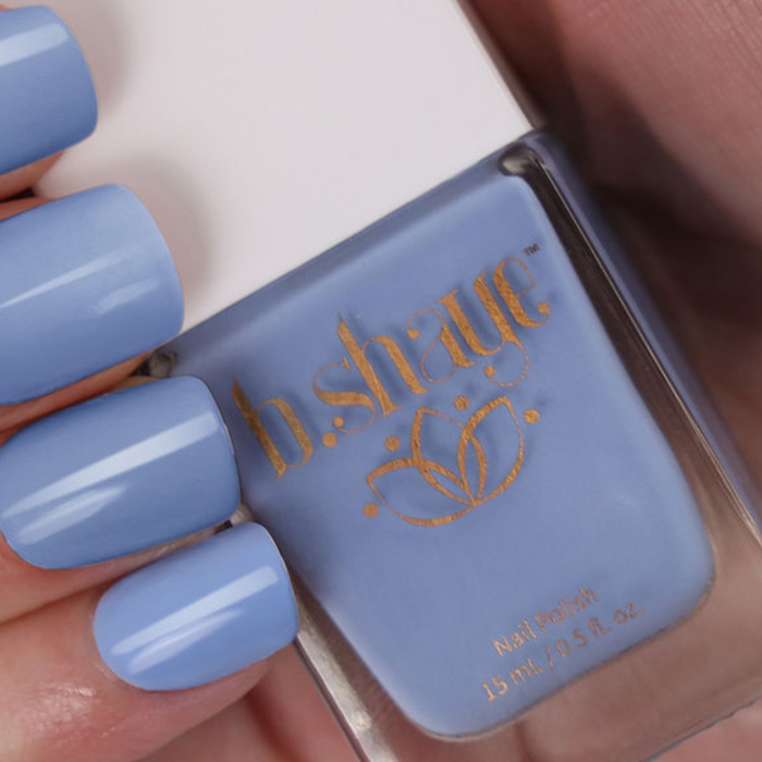 A hand with light blue nails holds a b.shaye nail polish bottle, adorned in light blue with gold floral design and promoting vegan, cruelty-free ingredients.