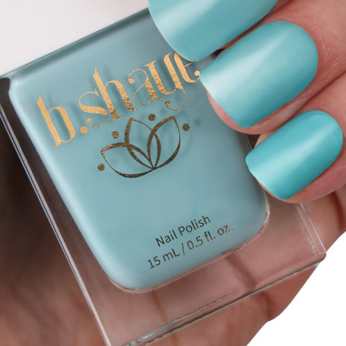 A hand with teal nails holds a 15 mL bottle of non-toxic b.shaye nail polish labeled in gold, representing clean beauty with its vibrant, vegan, and cruelty-free formula.