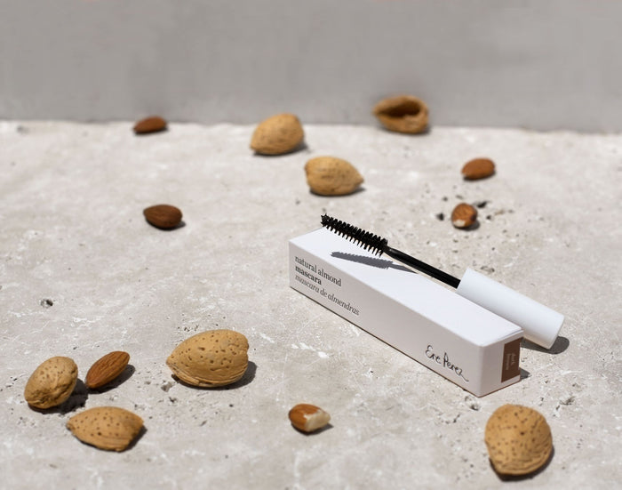 The Ere Perez Natural Almond Mascara, infused with nourishing almond oil for a smudge-proof finish, sits in its white box on a textured surface surrounded by almonds and their shells under soft natural light.