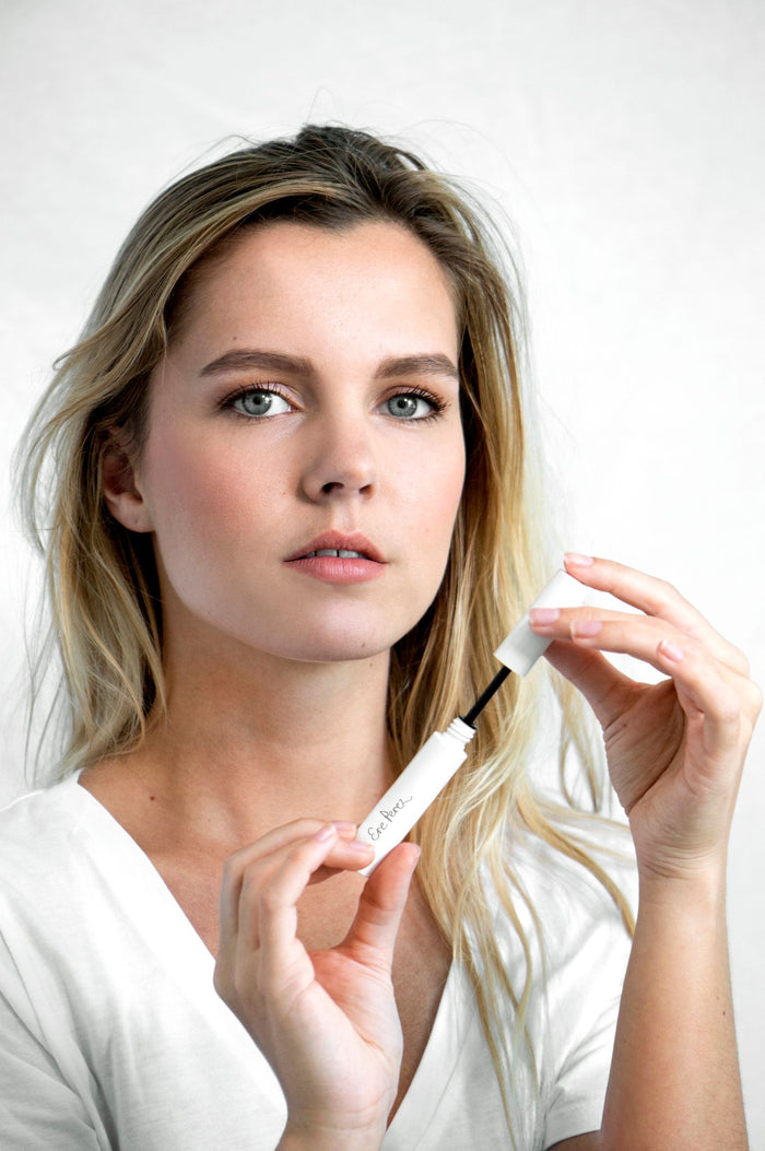 A person with long blonde hair holds an open tube of Ere Perezs Natural Almond Mascara, known for its smudge-proof formula and almond oil, wand poised against a light background, while wearing a white shirt.