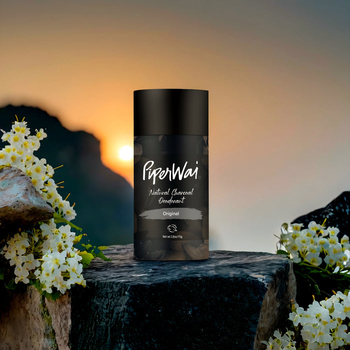 A Natural Deodorant Stick by PiperWai, infused with activated charcoal, rests on a rocky surface surrounded by blooming white flowers. The sky, aglow with sunrise or sunset hues, creates a serene backdrop for this natural deodorant masterpiece.
