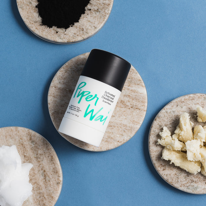 A PiperWai Natural Deodorant Stick, featuring activated charcoal, is placed on a round stone slab. Around it are two slabs displaying black powder, white chunks, and a creamy texture with a blue background.