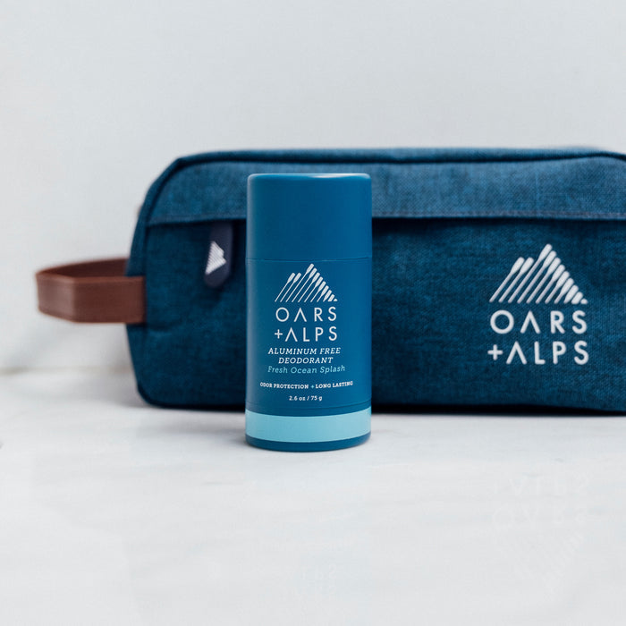 An Oars + Alps Natural Deodorant in blue stands on a white surface before a matching travel bag featuring the brand’s logo. The setting has a clean, minimalistic background.