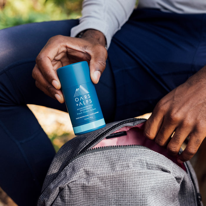 Someone puts an Oars + Alps Natural Deodorant stick into a gray backpack with a pink lining while sitting outside in casual athletic wear.