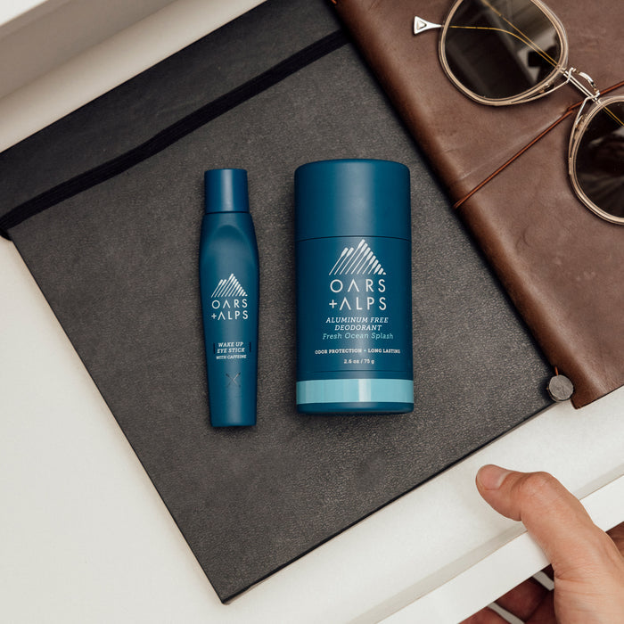 A hand reaches toward a black notebook topped with two Oars + Alps Natural Deodorant sticks, set beside brown sunglasses and a leather item on a white surface.