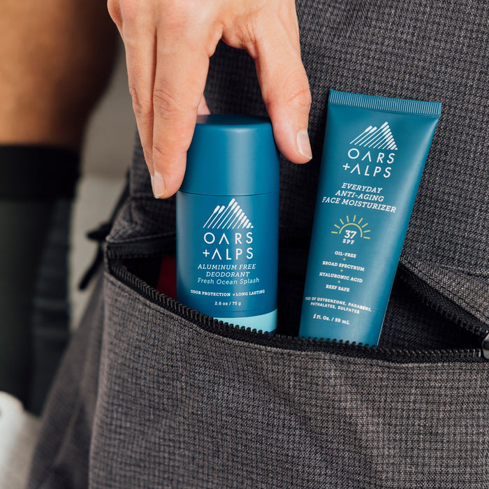 A hand places an Oars + Alps Natural Deodorant and face moisturizer—both blue with white lettering—into the front pocket of a grey bag. This odor-fighting pair is a Mens Health Editors Choice.