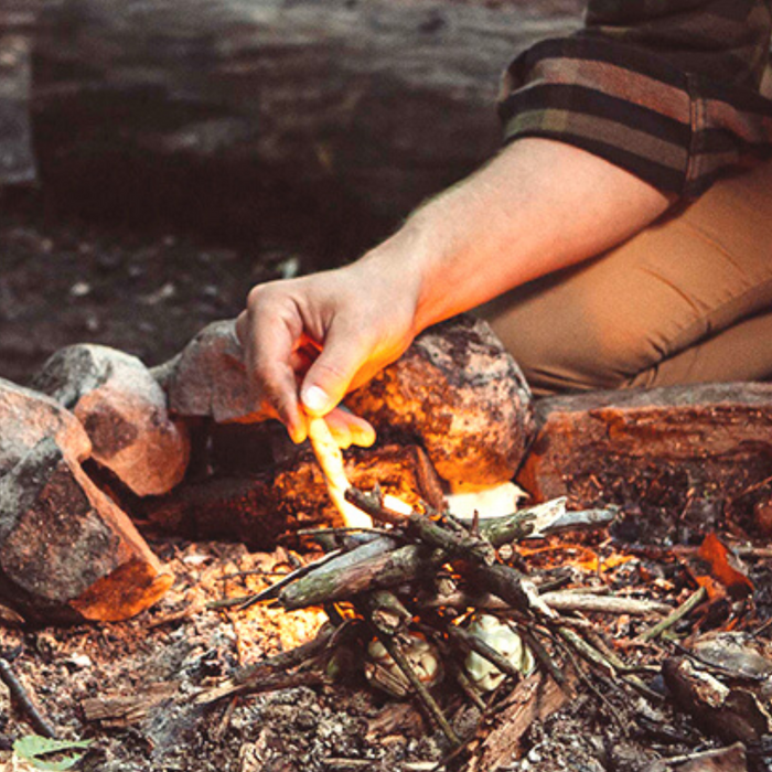 Wearing a plaid shirt, a person crouches to use ReKindle Fire Starters by Bees Wrap to ignite a small campfire made of twigs and leaves. Surrounded by stones, the fire flickers warmly in the natural outdoor setting.