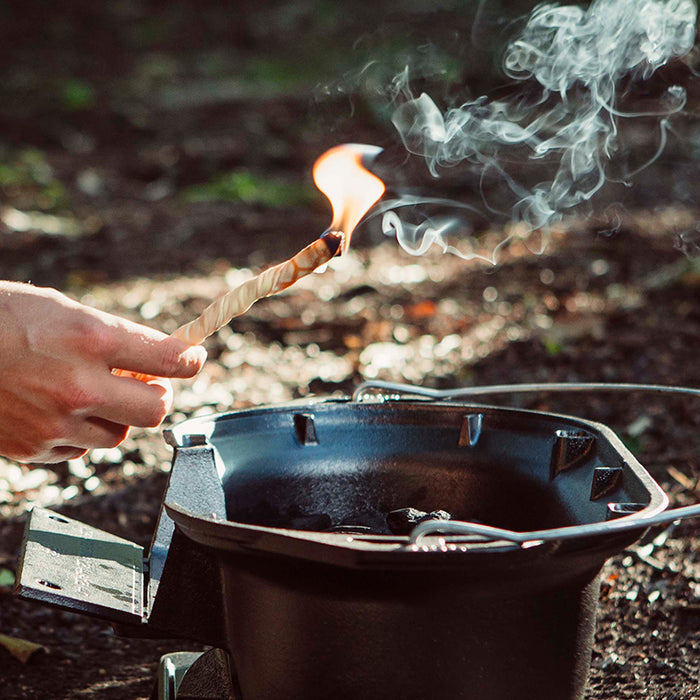 Someone uses a ReKindle fire starter by Bees Wrap to light a twisted wooden stick above a small black outdoor grill. Smoke rises, evoking the experience of starting campfires. The background features blurred natural scenery with dappled sunlight on the ground.