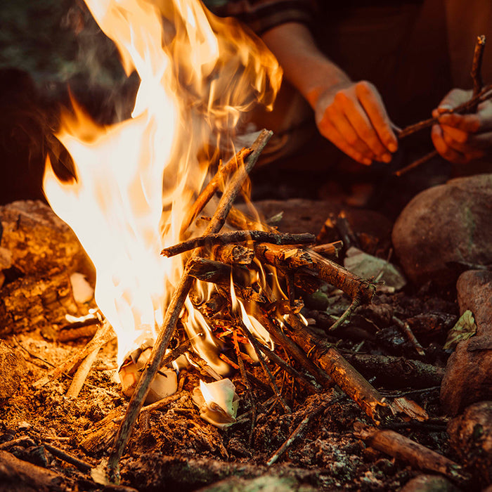 Close-up of a campfire with bright orange flames surrounded by rocks, as someone uses ReKindle fire starters from Bees Wrap to add small twigs, their hands partially visible. The setting suggests an outdoor, possibly evening atmosphere.
