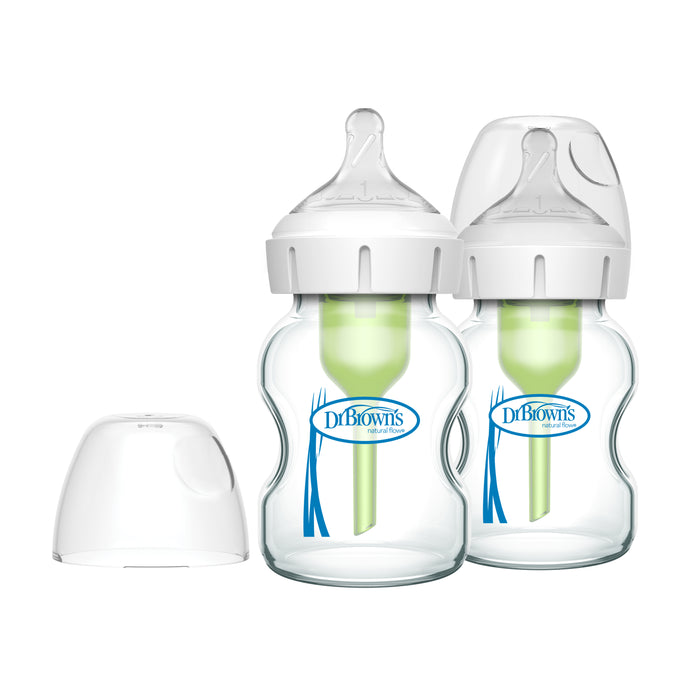 Two clear Dr. Browns Natural Flow Options+ wide-neck glass baby bottles come with green anti-colic vent systems, white silicone nipples, and extra caps. Designed for easy filling and cleaning, the trusted brand is labeled in blue text, ensuring confidence for parents.