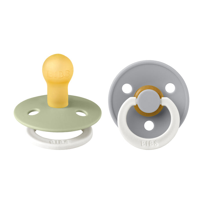 Two BIBS pacifiers: one olive green/mustard yellow, the other gray with a white ring handle—each with a round, flat shield, two vent holes, and embossed handles. Made from BPA-free natural latex rubber for seamless breastfeeding; available as Glow in the Dark Pacifier, Size 1 (0-6 Months).